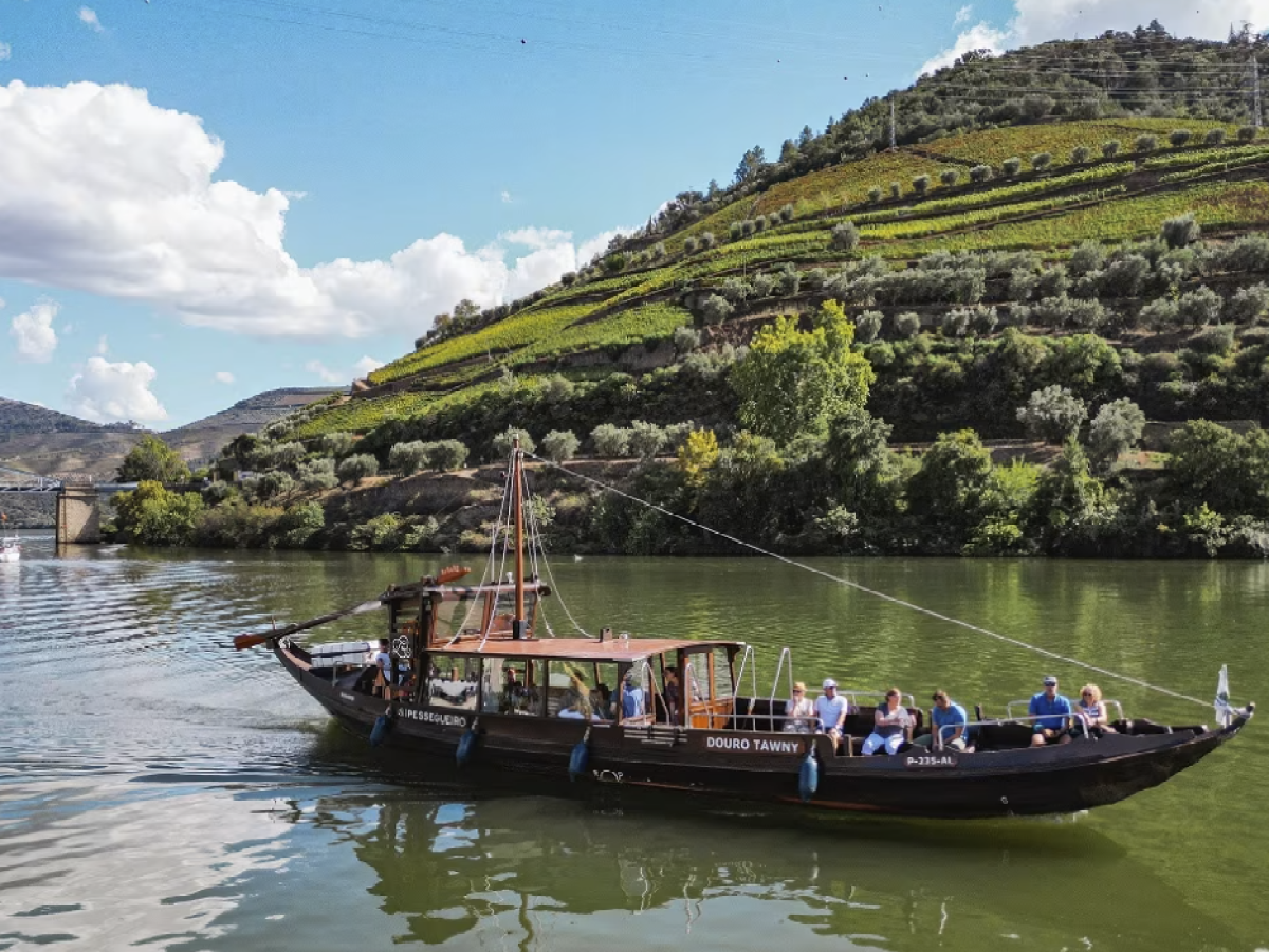 A traditional boat with tourists on a river, green hills in the background under a blue sky with clouds.