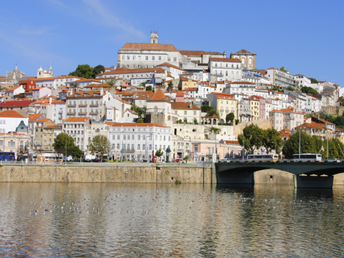 Hillside cityscape with colorful buildings, a bridge, and a river in the foreground under a clear blue sky.