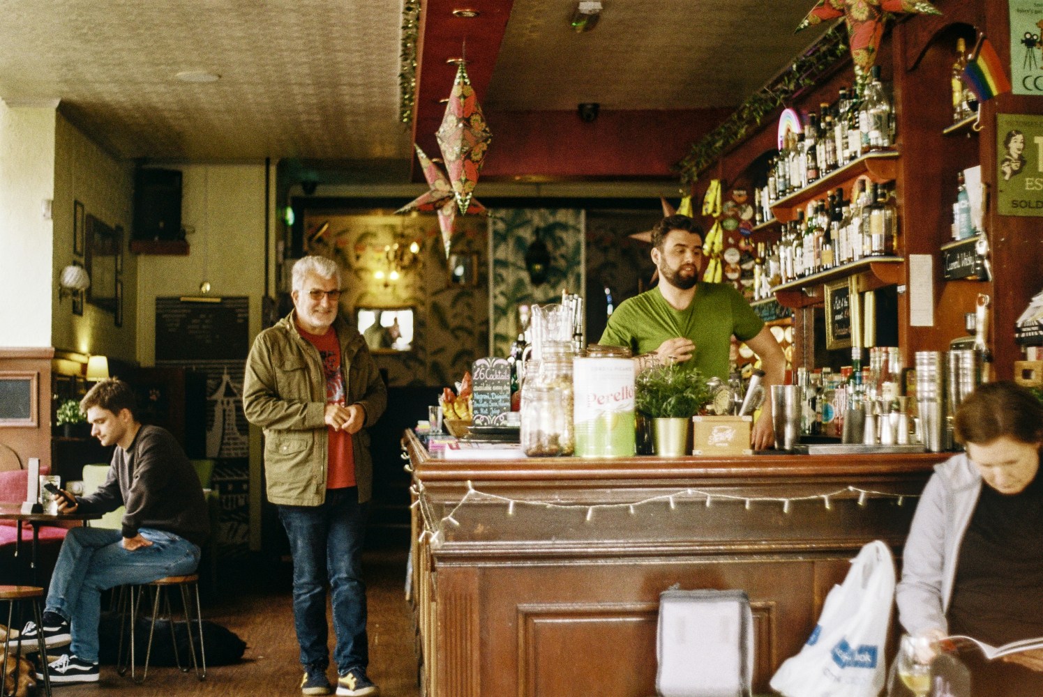 Bar interior with people, bartender in green shirt behind the counter, and a seated man using a phone.