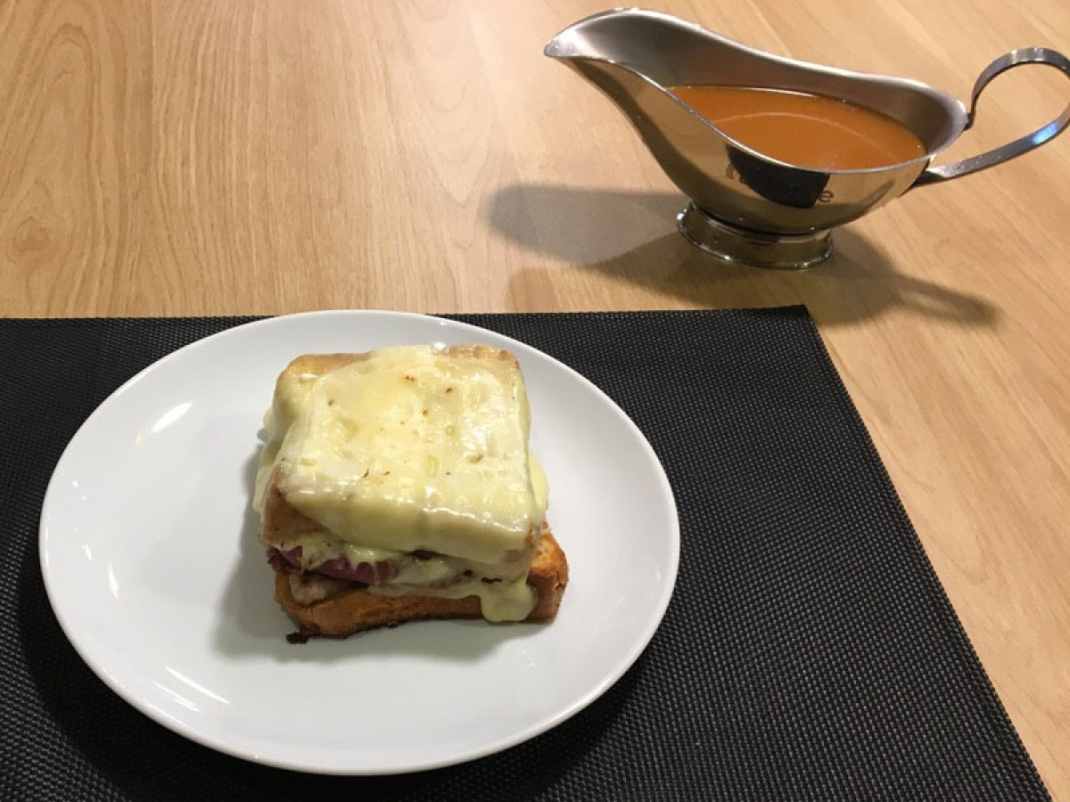 Cheese-topped sandwich on a plate with gravy boat on wooden table.