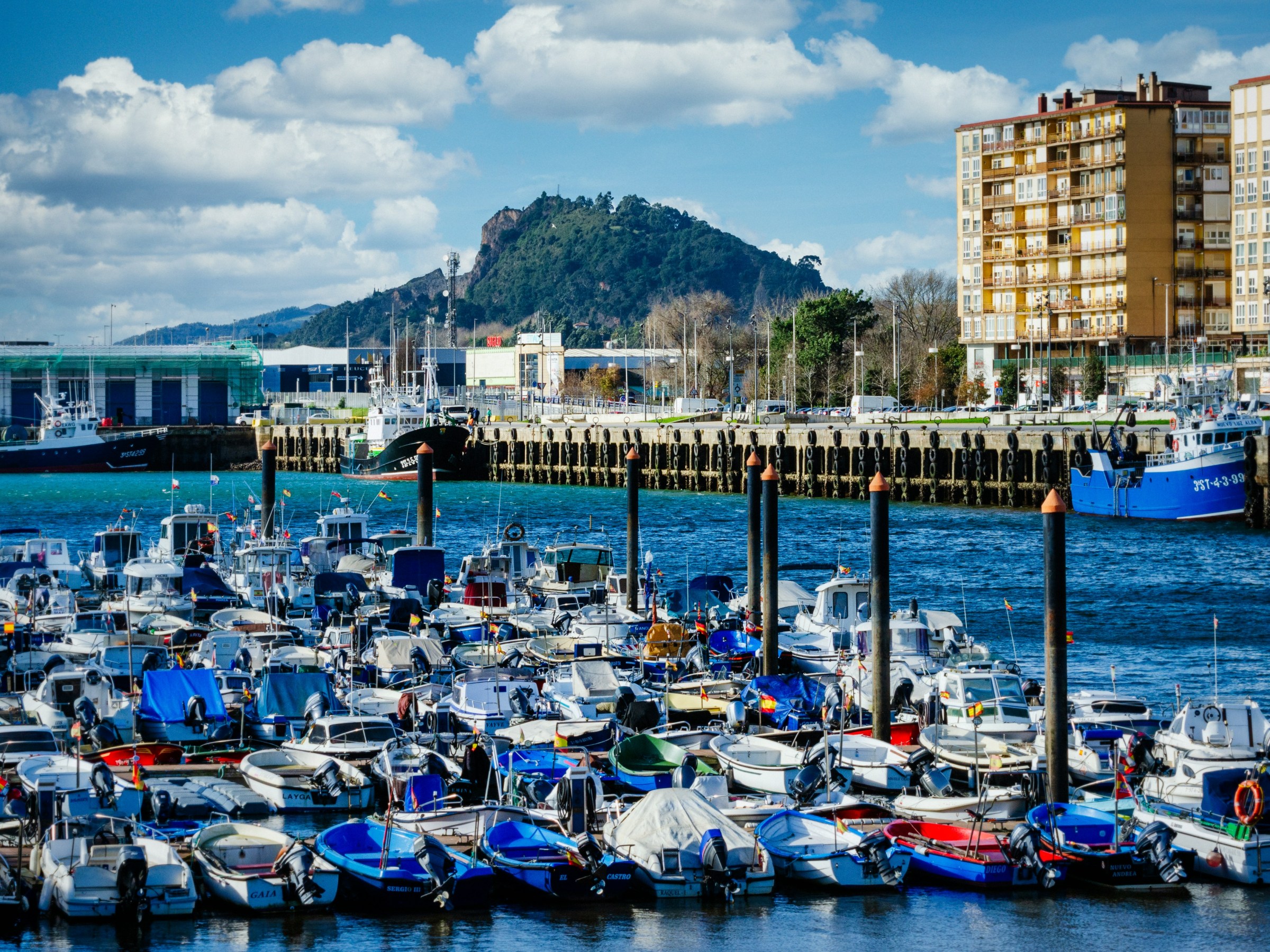 Boats docked in a marina with buildings and a hill in the background under a partly cloudy sky.