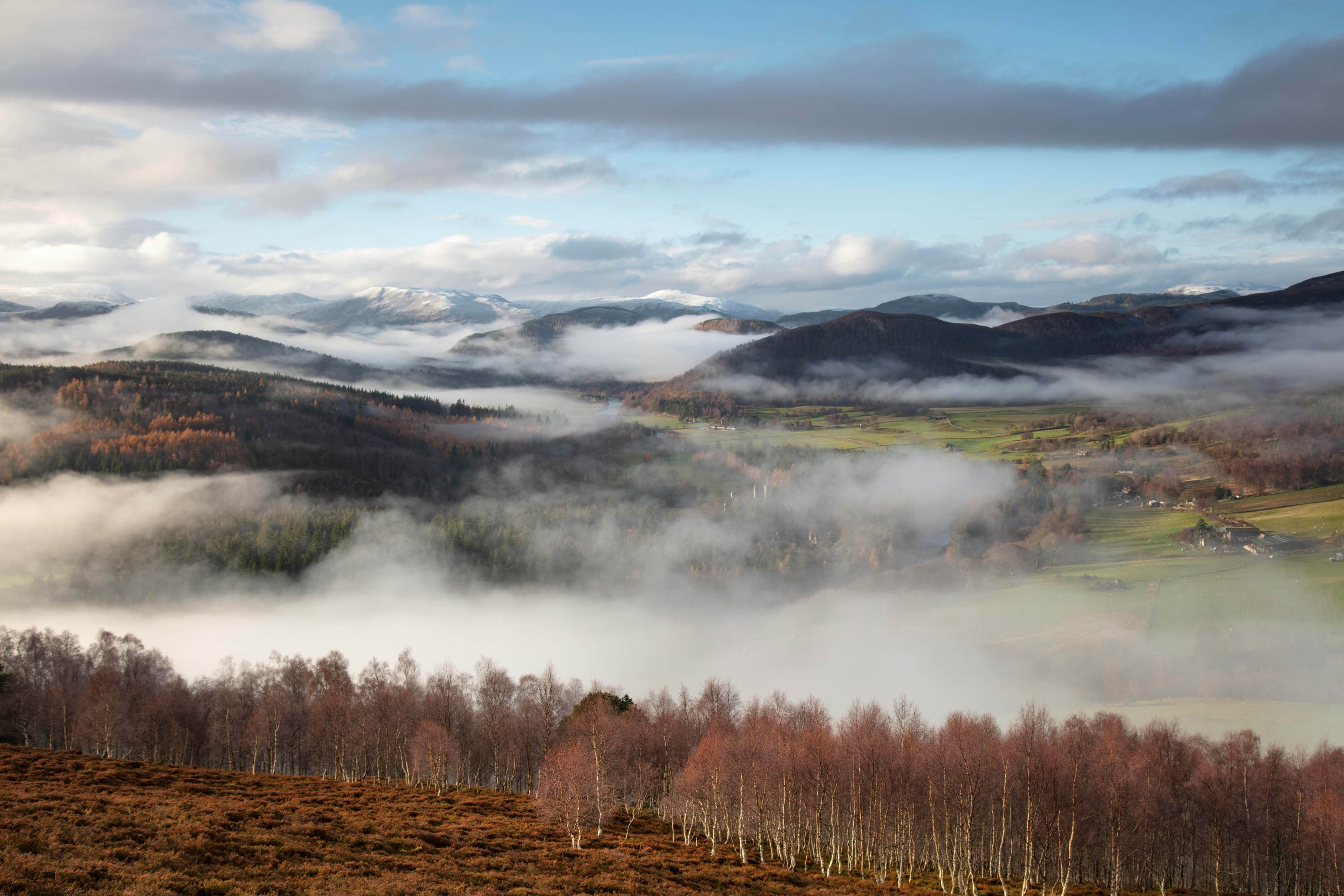 Fog over a valley with hills, trees, and distant mountains under a blue sky.
