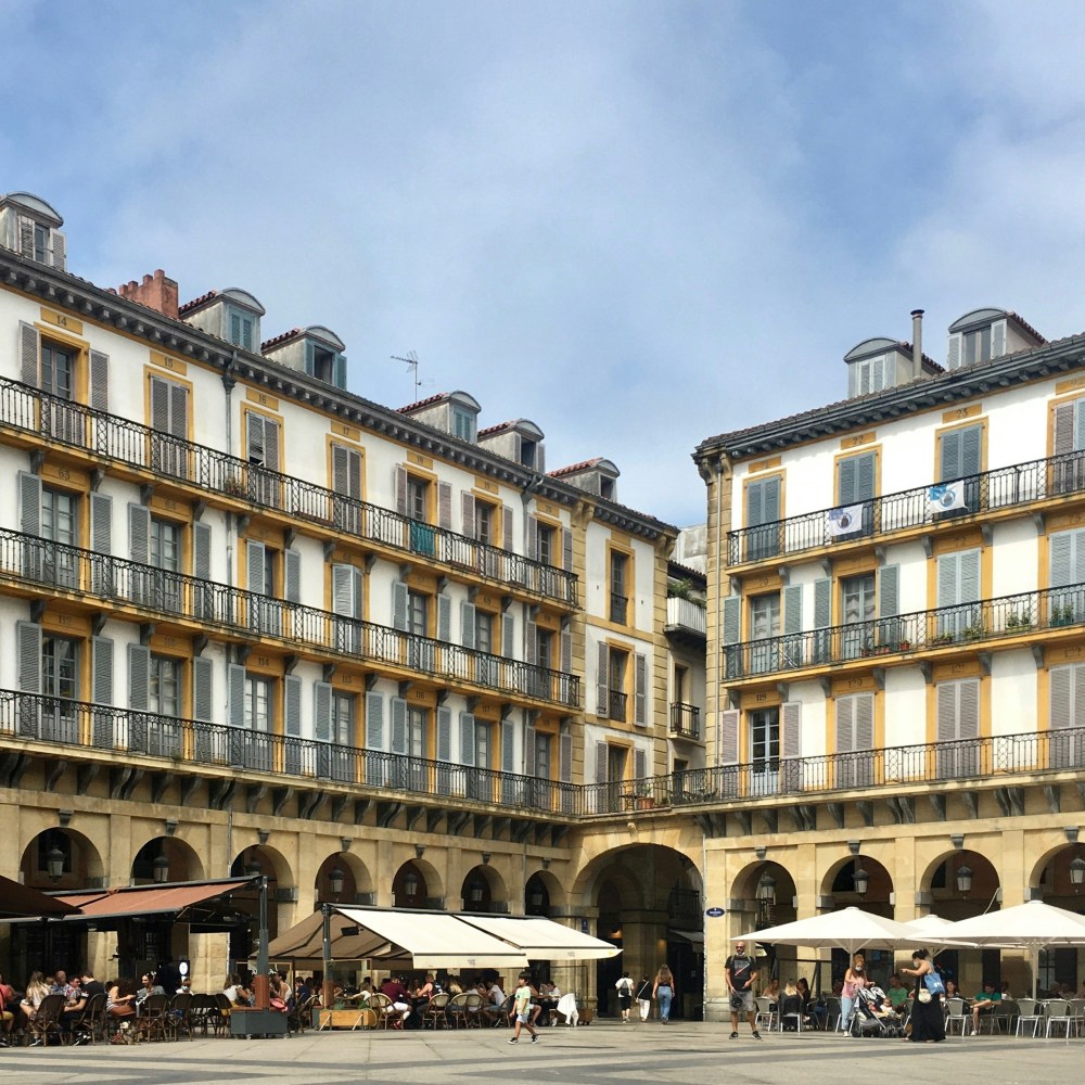 People dining outside a historic building with yellow balconies and gray shutters in a sunny plaza.