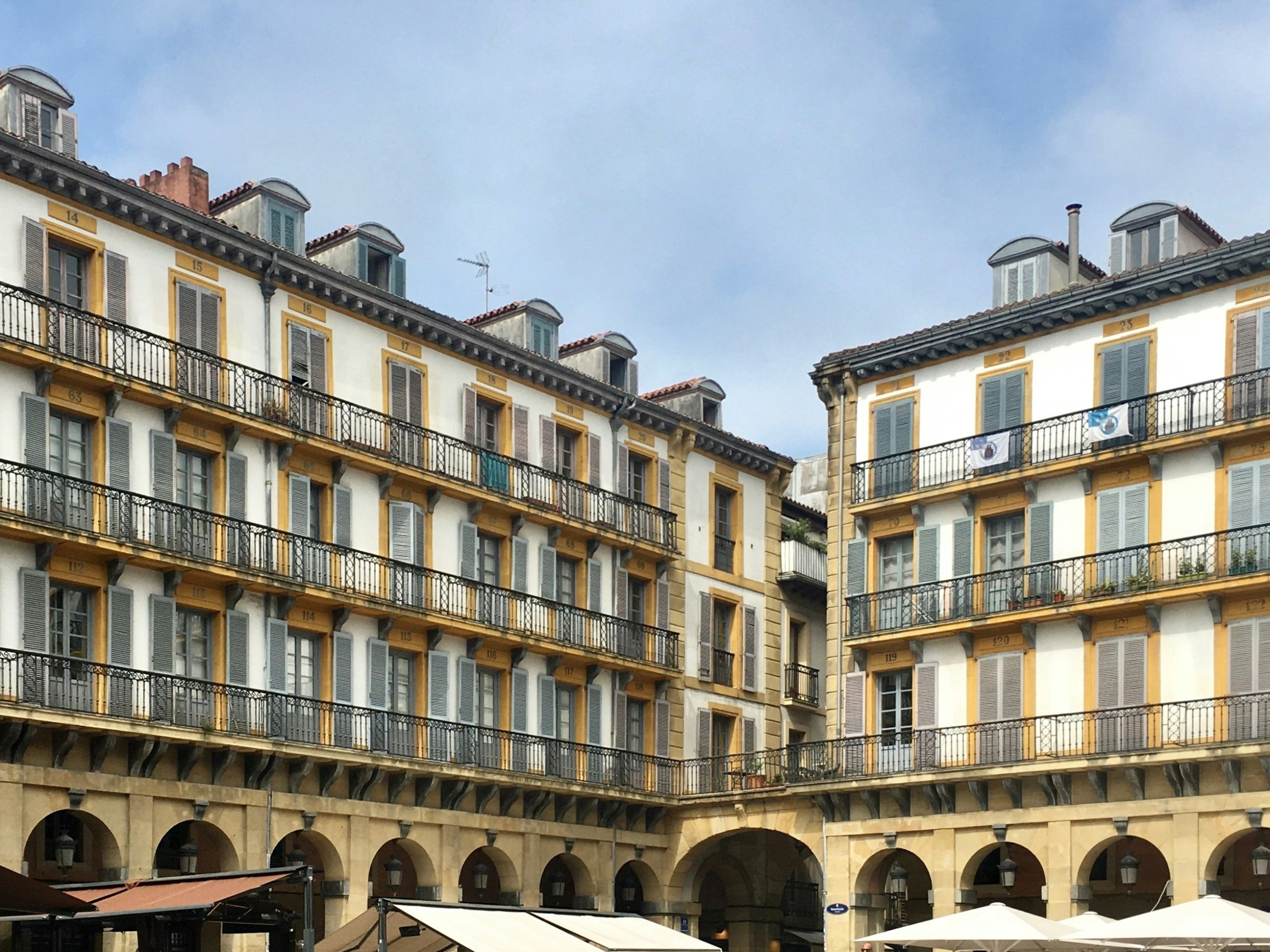 People dining outside a historic building with yellow balconies and gray shutters in a sunny plaza.