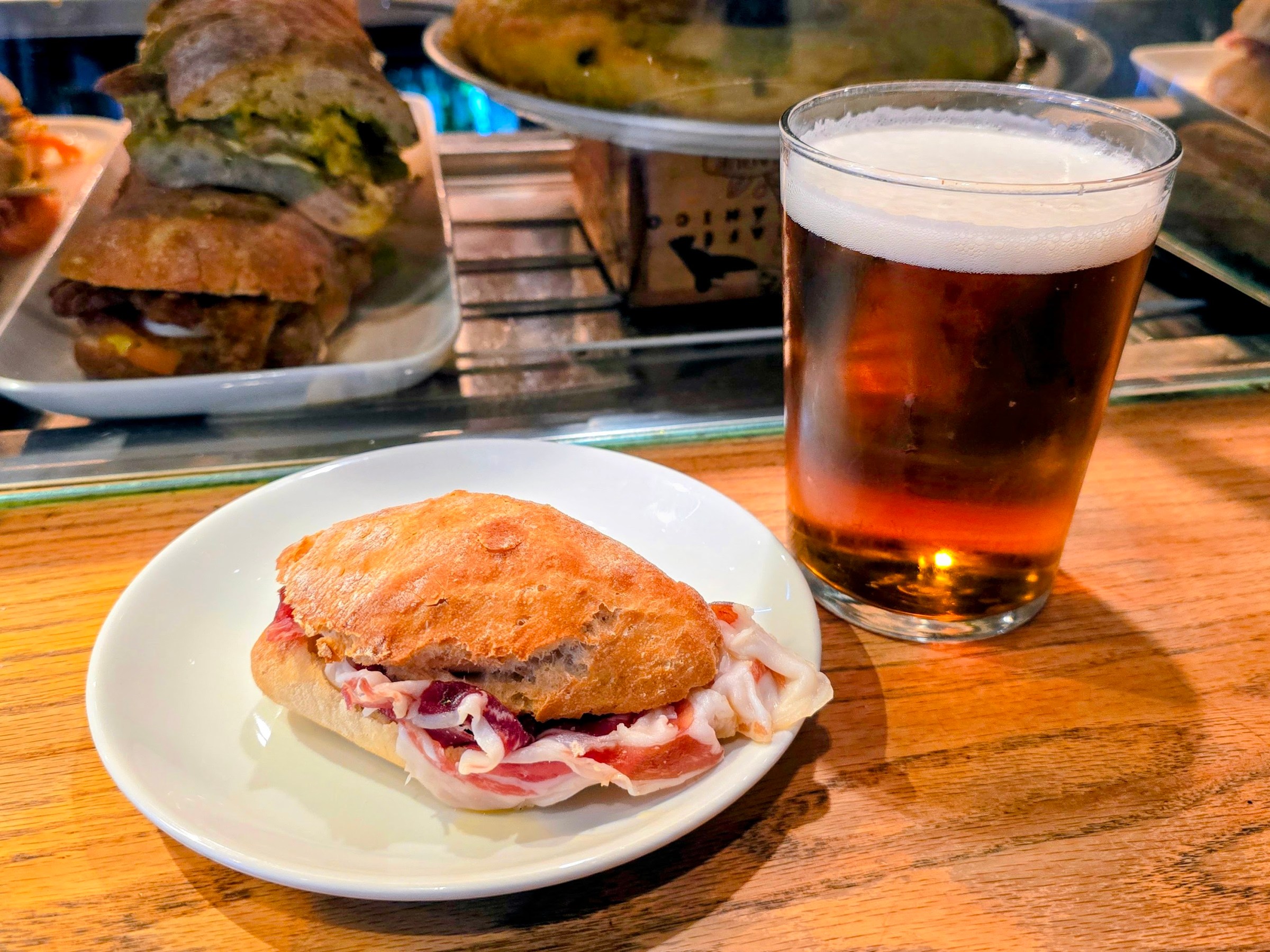 Small sandwich on a plate next to a glass of beer on a wooden table.
