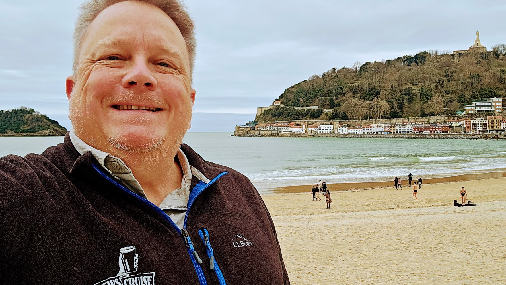 Smiling person taking a selfie on a beach with hills and buildings in the background.
