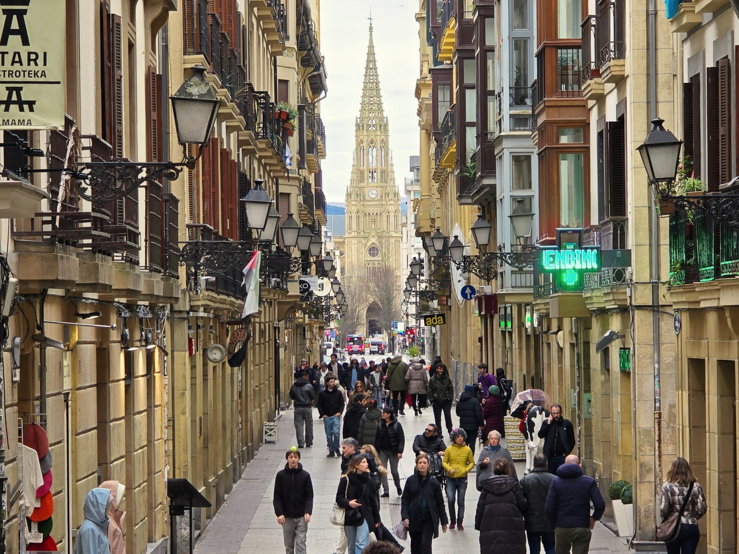 Crowded narrow street lined with buildings, leading to a cathedral tower in the background.