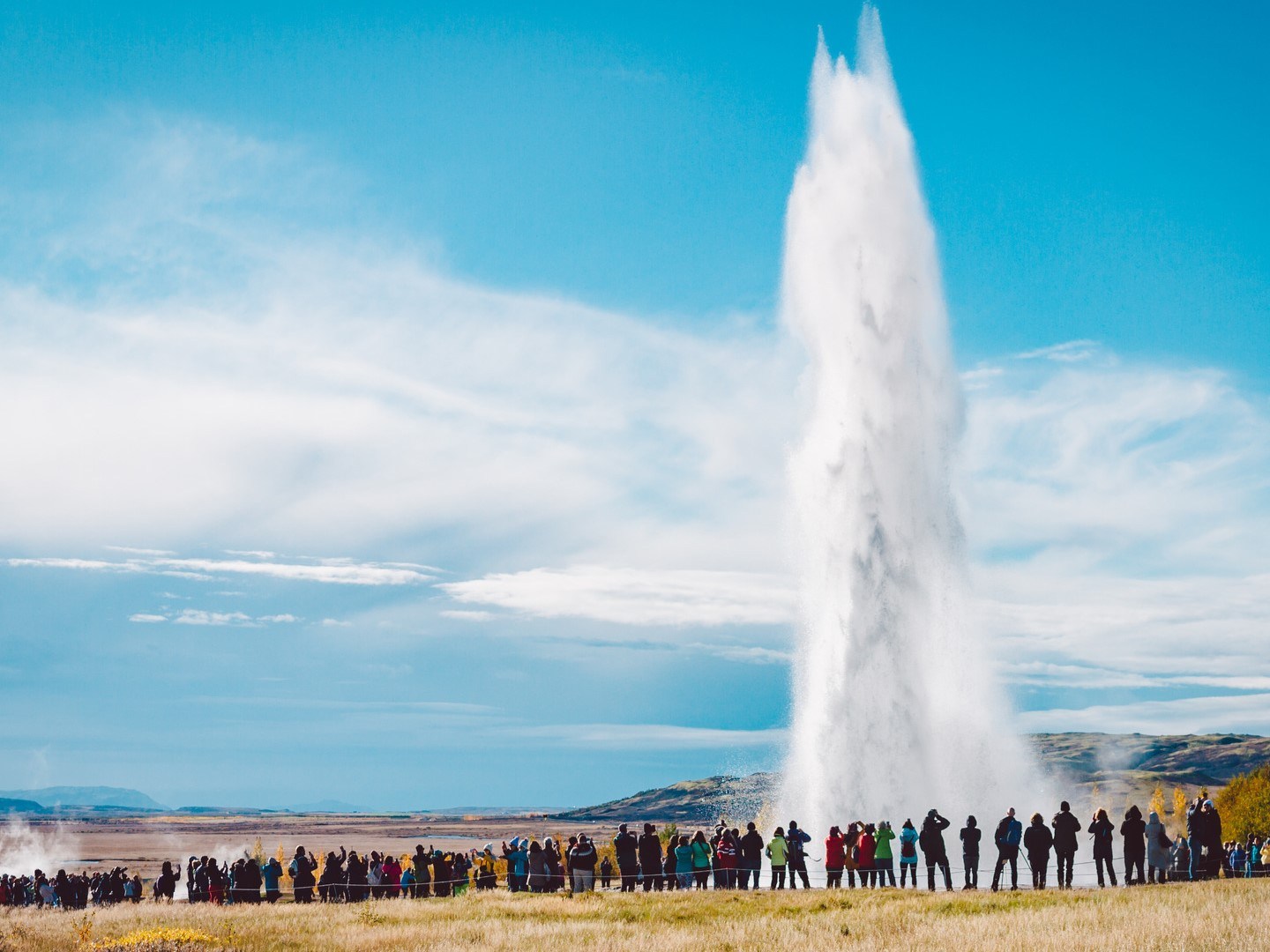 Crowd watching a large geyser erupt under a blue sky with clouds.
