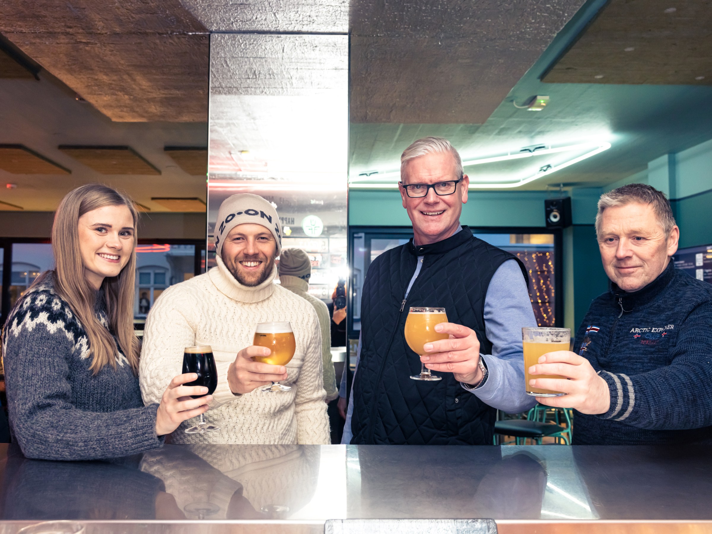 Four people at a bar, smiling and holding beer glasses, wearing warm clothing.