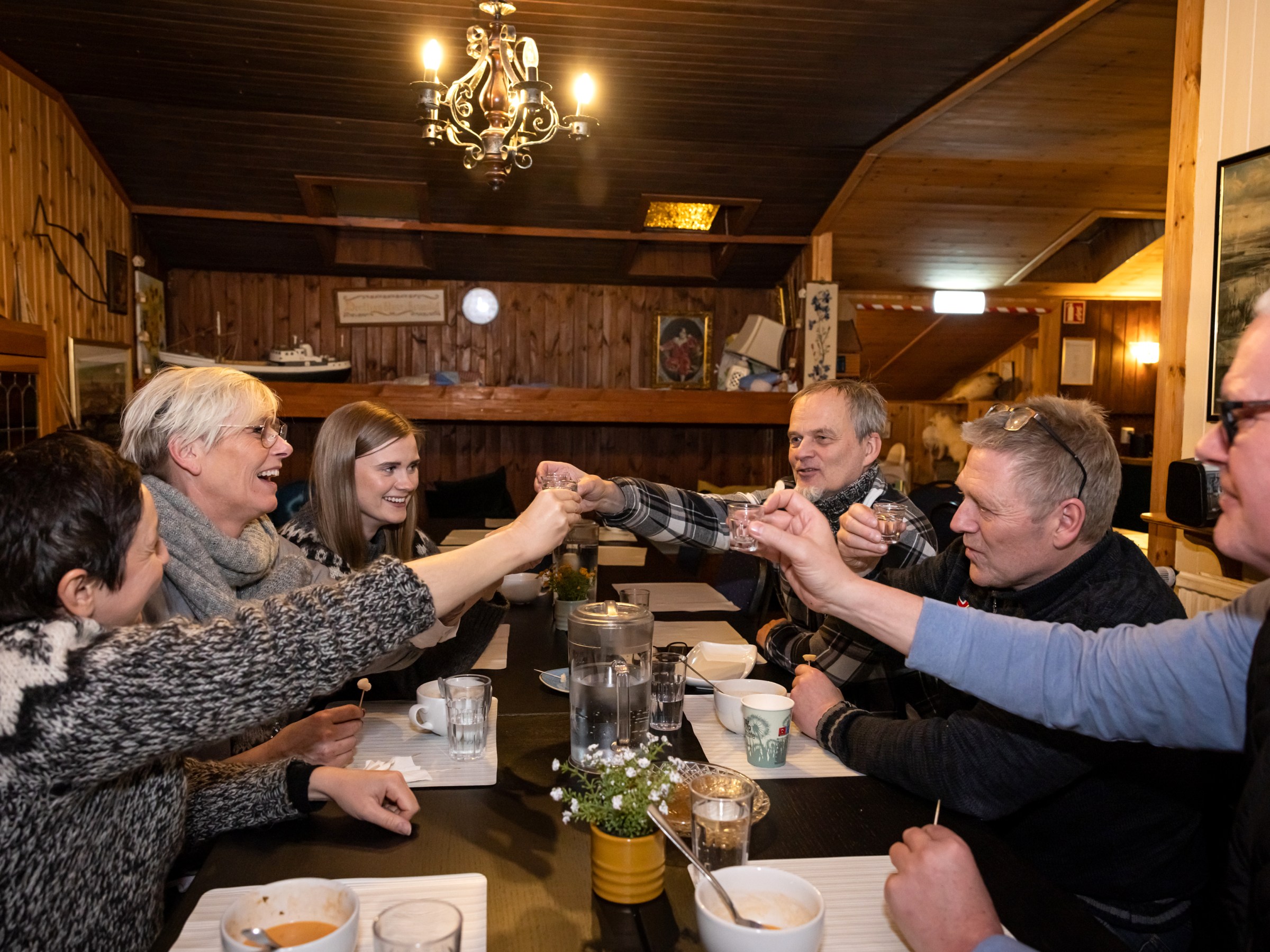 Group of six people sitting in a cozy room, toasting with small glasses over a dining table.