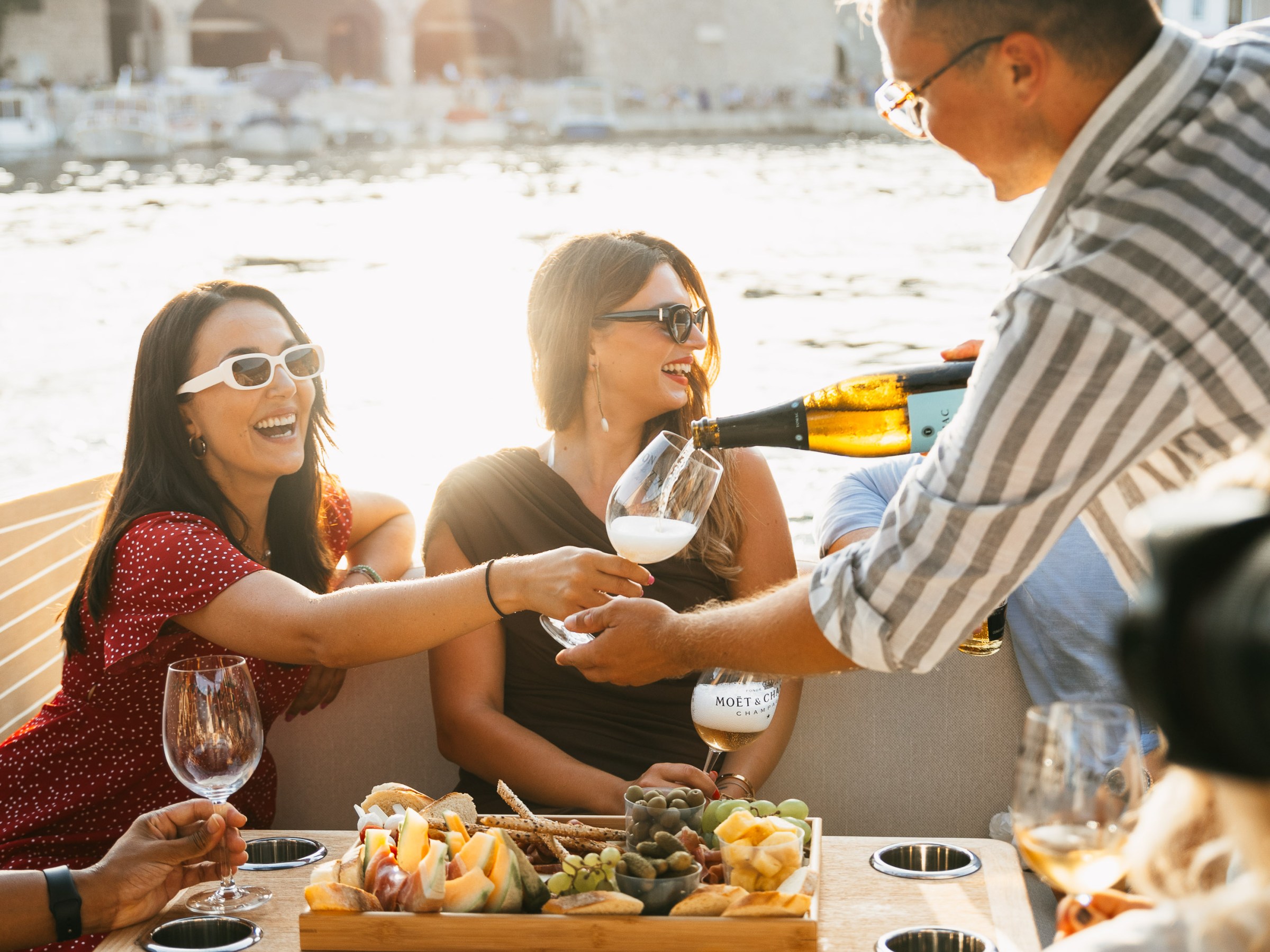 People enjoying a wine and fruit platter on a boat with a scenic view of water and buildings.