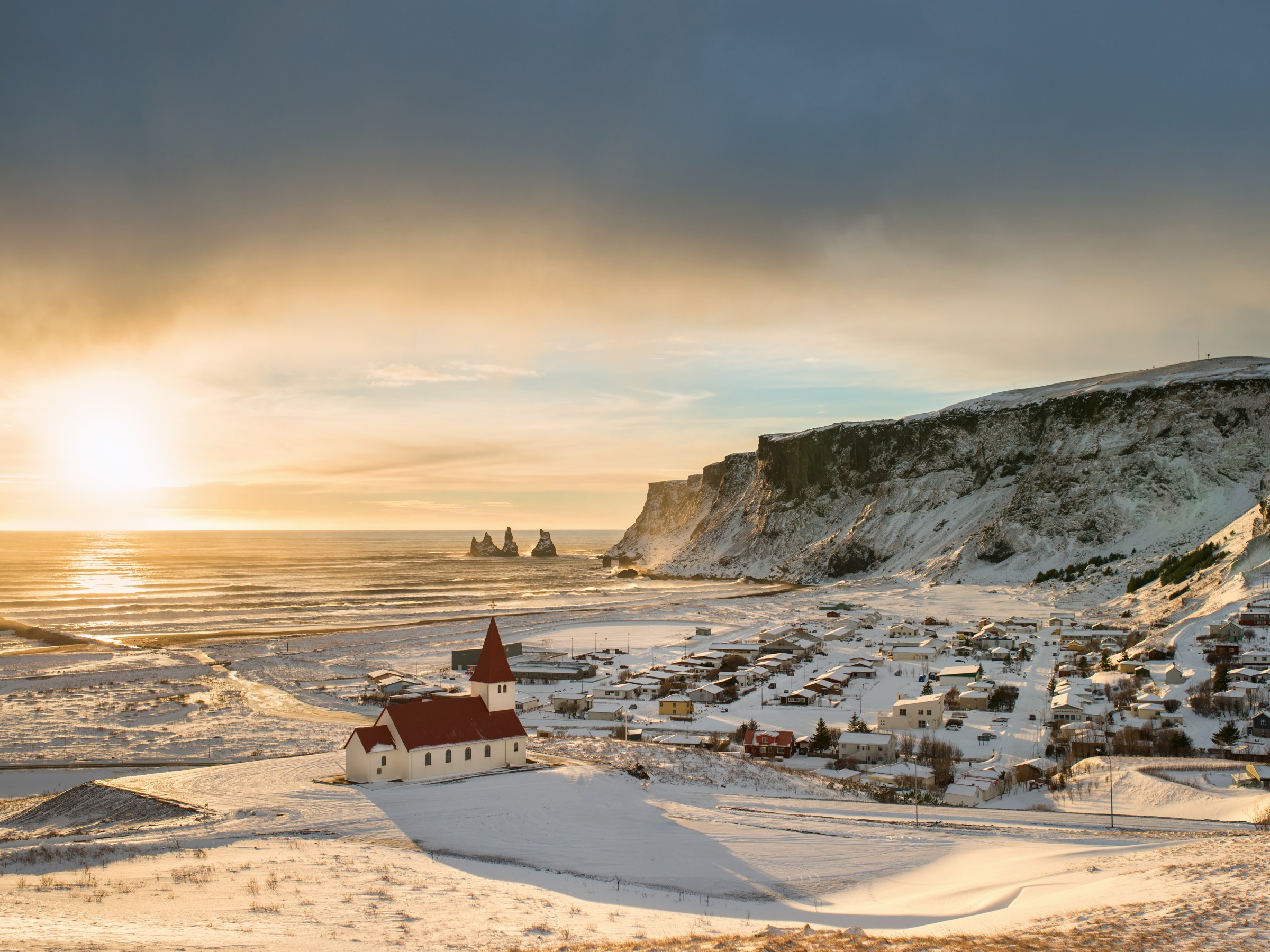 Snowy coastal town with a red-roofed church, cliffs, and ocean view at sunrise.