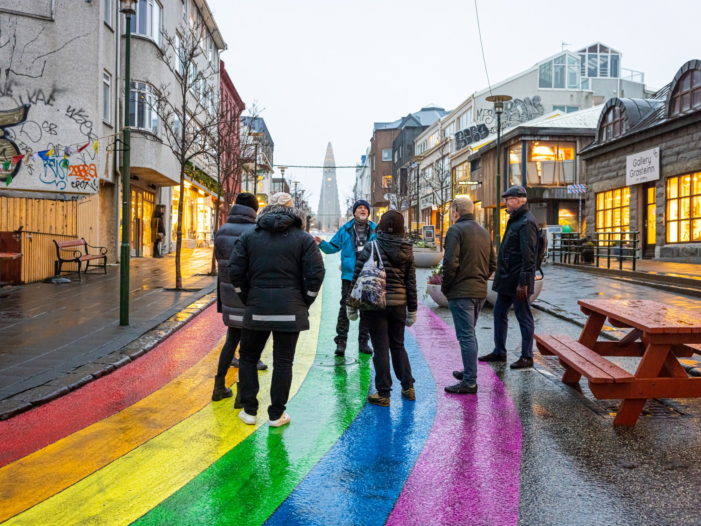 Group standing on rainbow-painted street in a city with graffiti and storefronts.