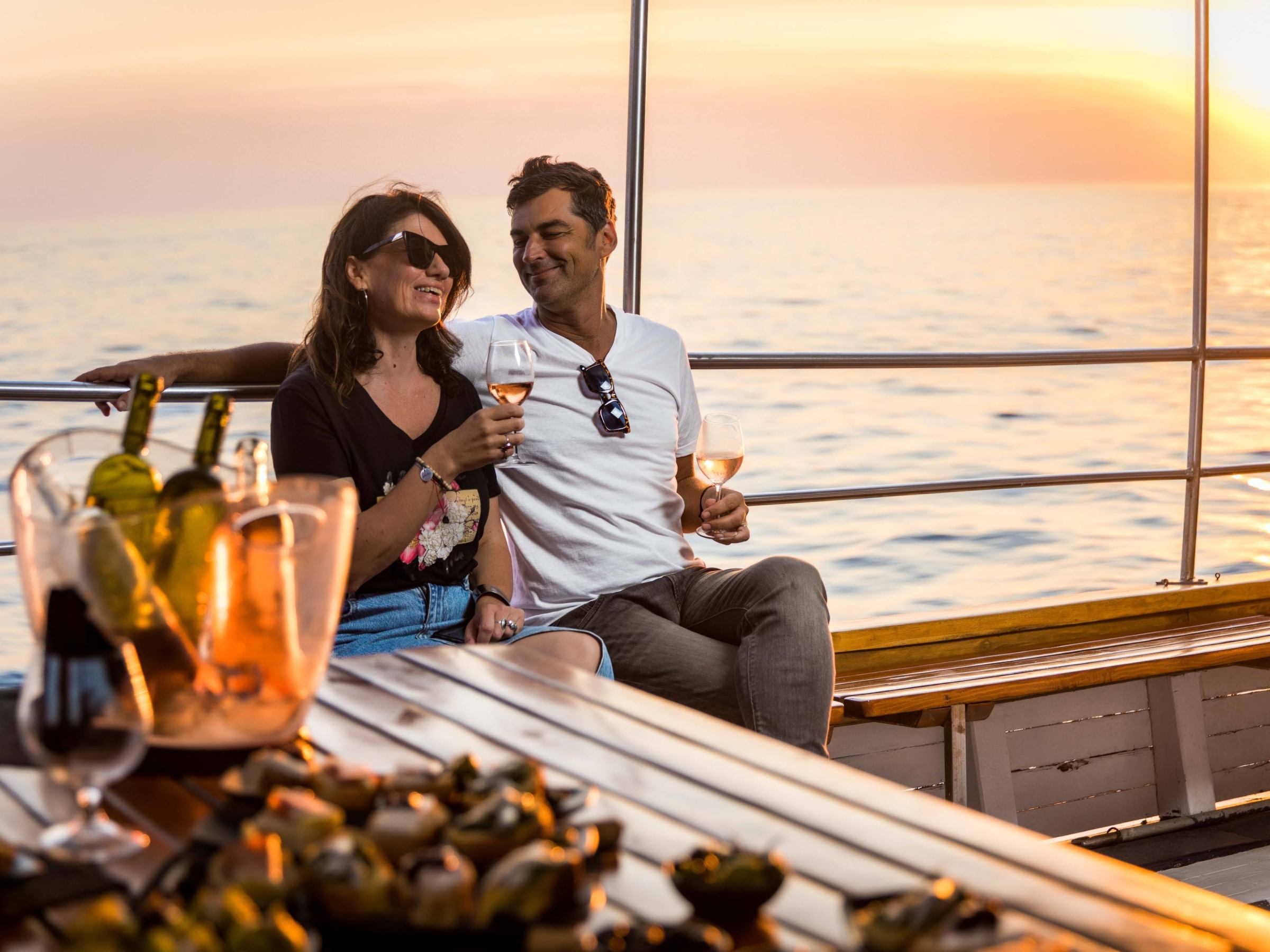 Couple enjoying drinks on a boat at sunset with wine bottles on the table.