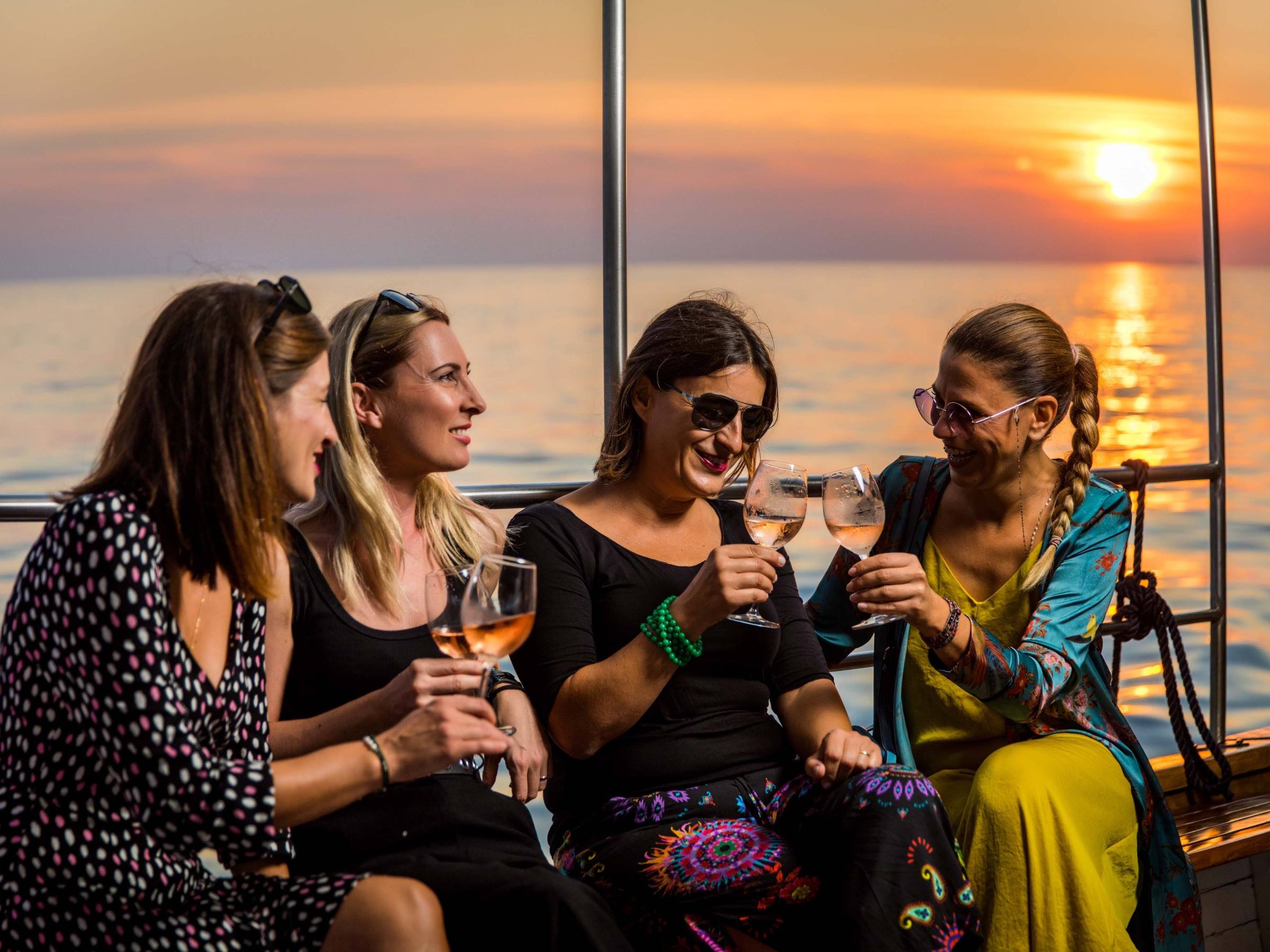 Four women on a boat toasting with wine glasses at sunset.