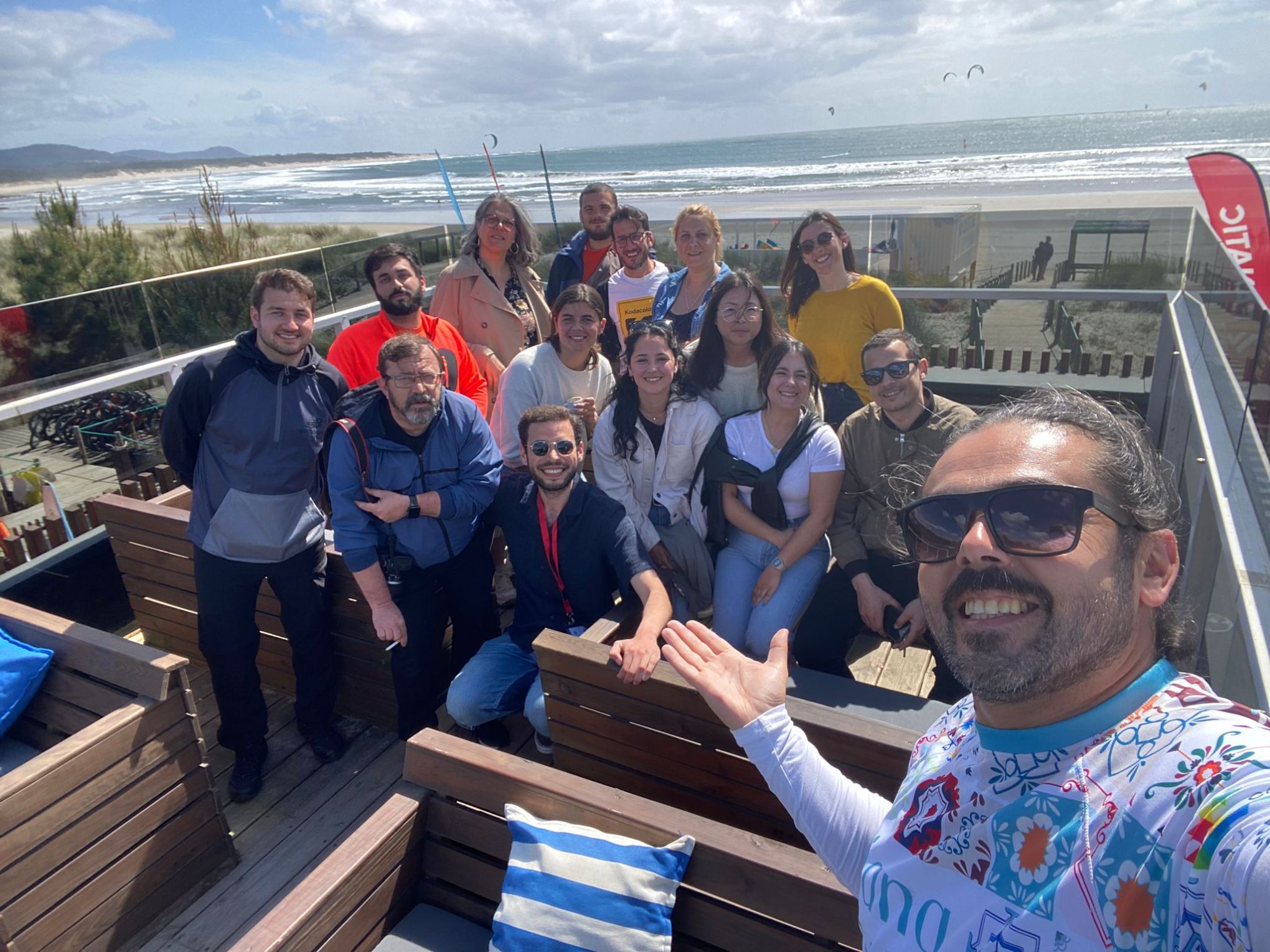 Group of people smiling on a wooden deck near the beach with waves and kite surfers in the background.