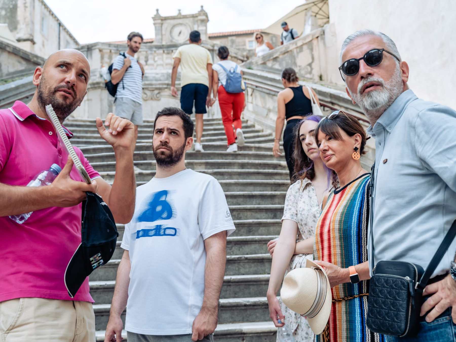 Group of people standing on stone steps, some looking forward, urban setting.