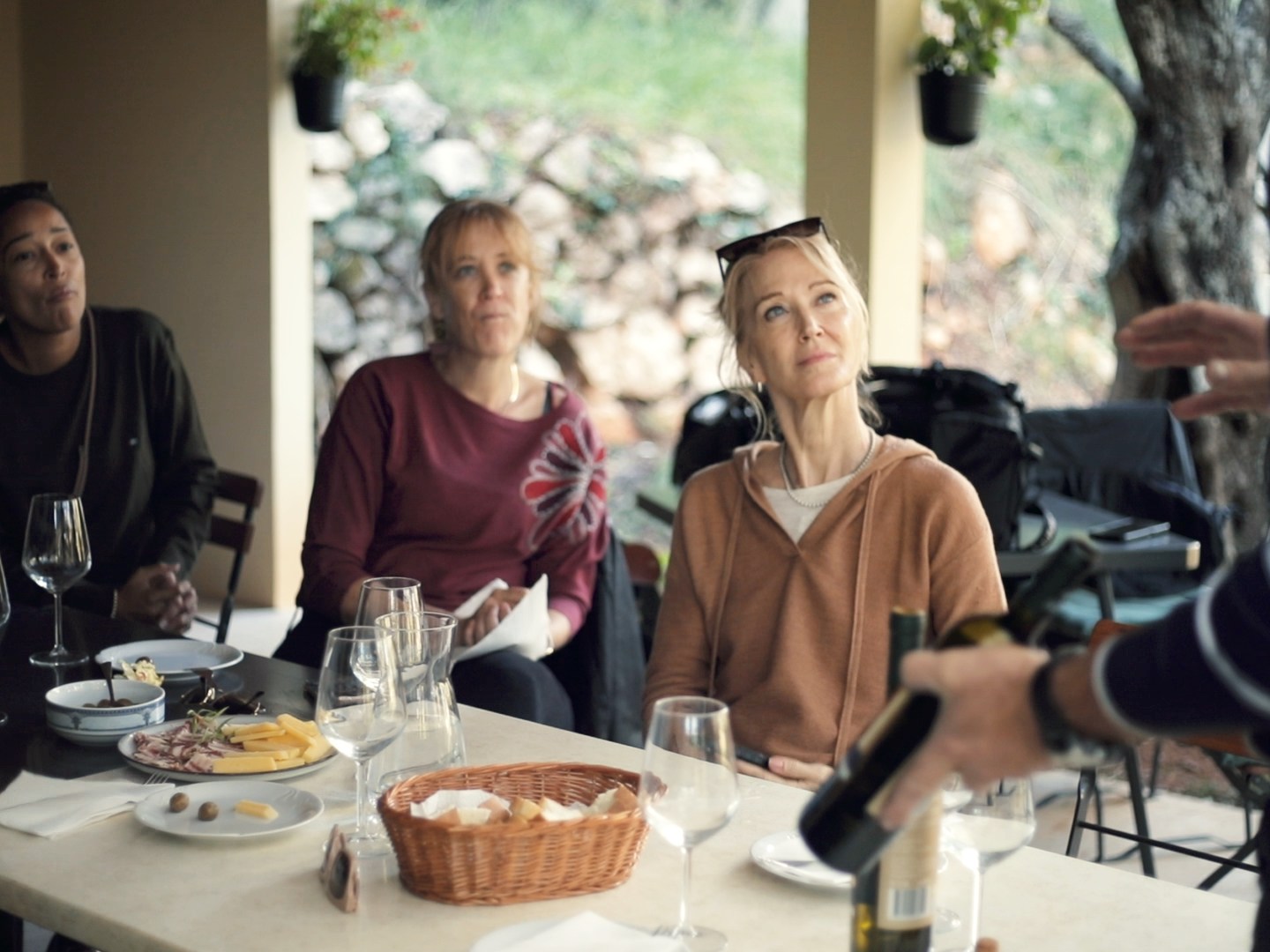 People sitting at a table with wine glasses and snacks, listening to a standing person holding a bottle.