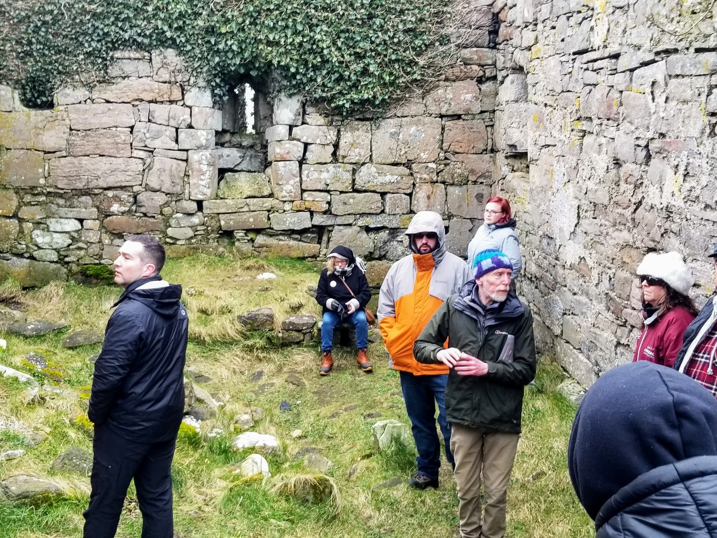 People in jackets explore stone ruins with grassy floor.