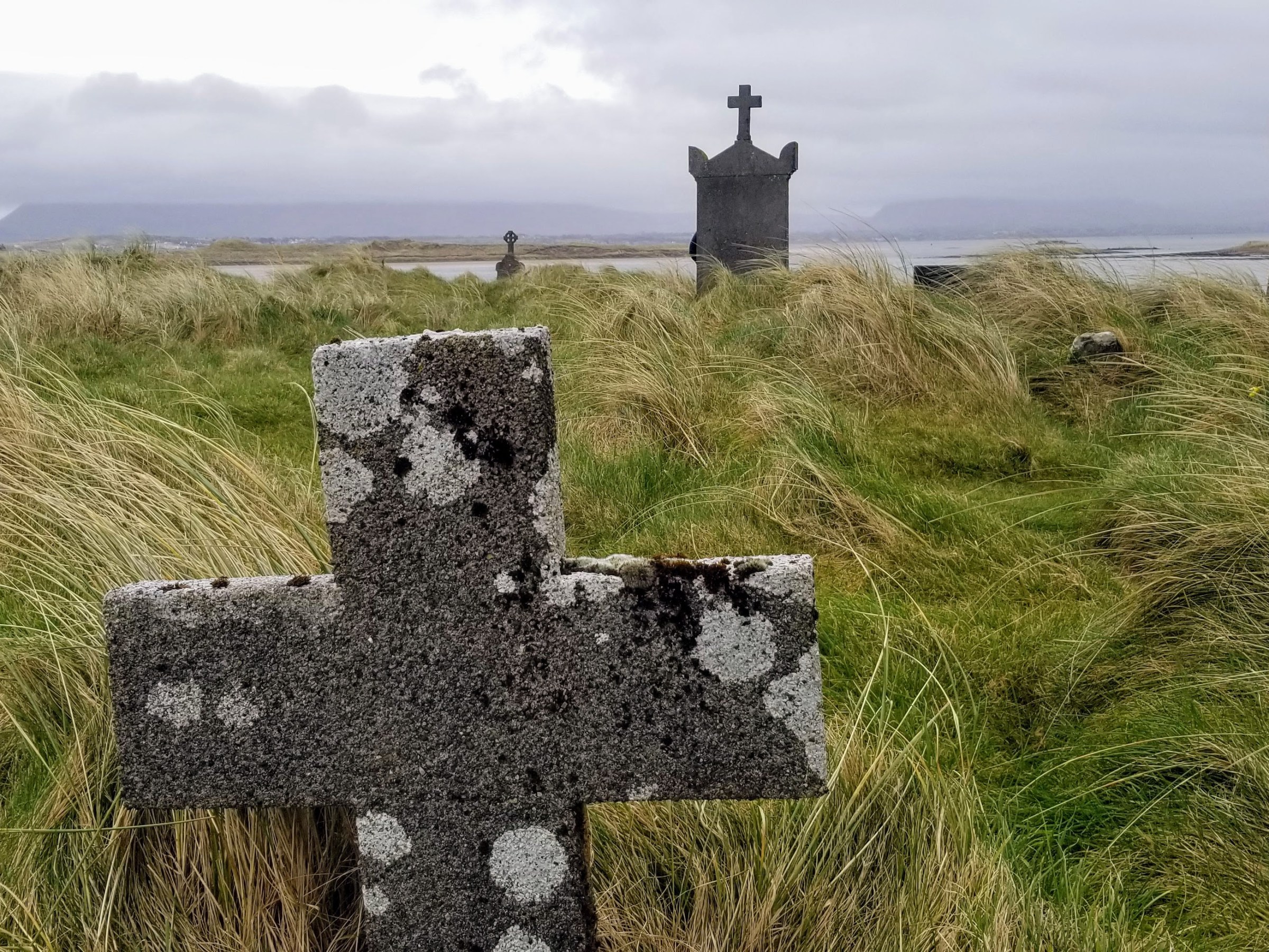 Stone crosses in a grassy, remote cemetery with cloudy sky.