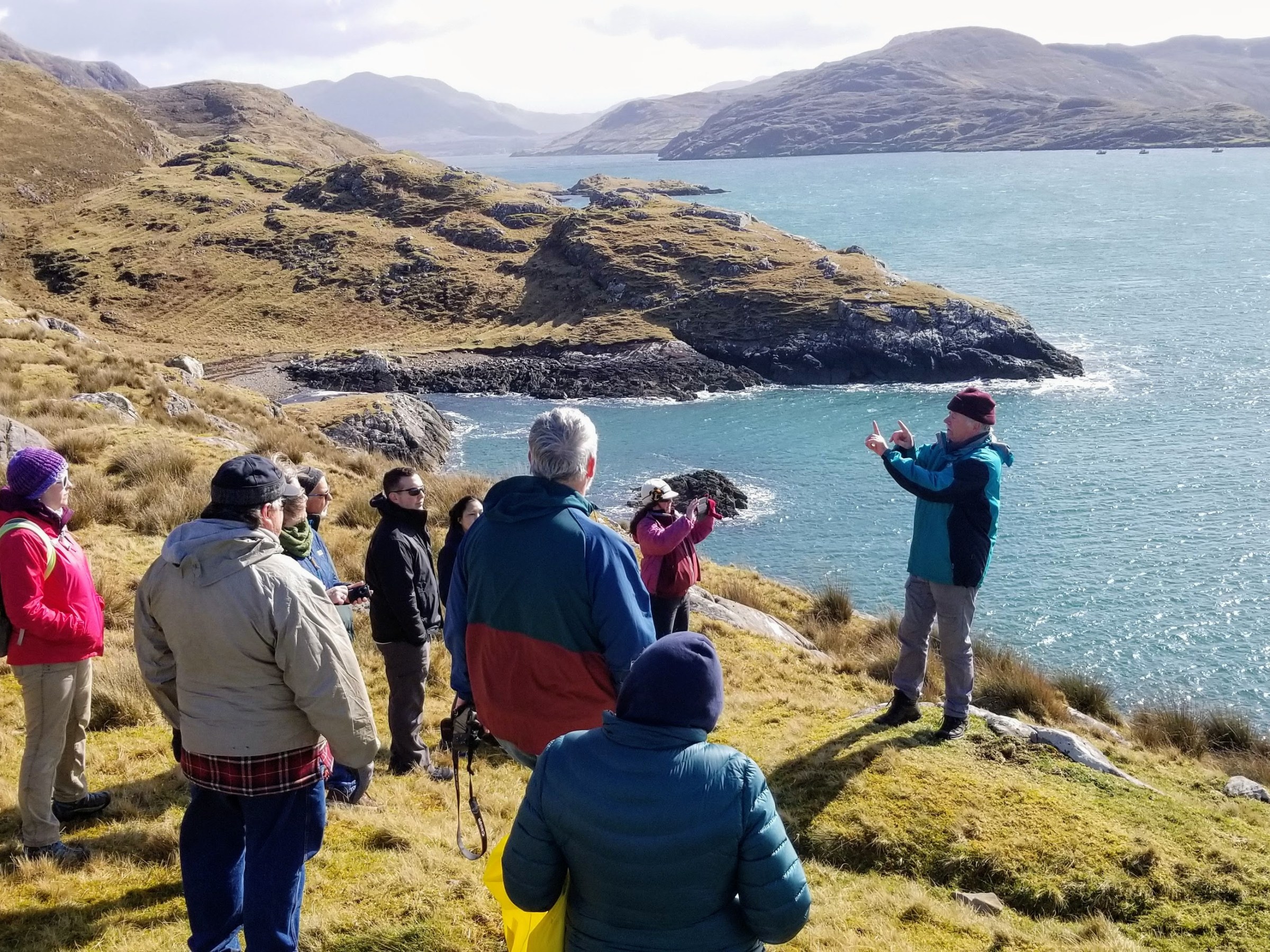 A group of people stand on a grassy hill overlooking a scenic body of water and mountains.