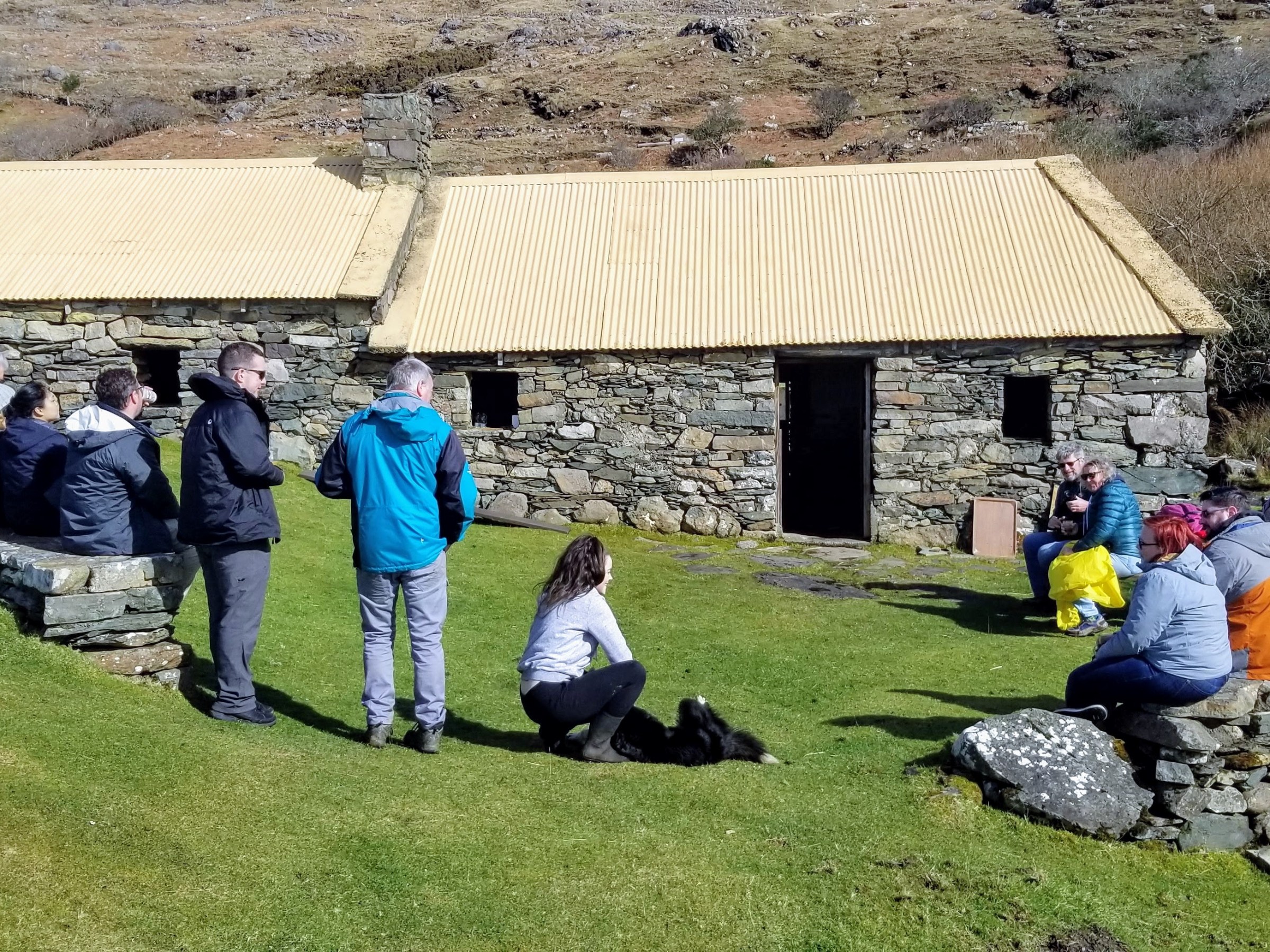 Group gathered outside stone building with yellow roof, seated on grass and stone wall.