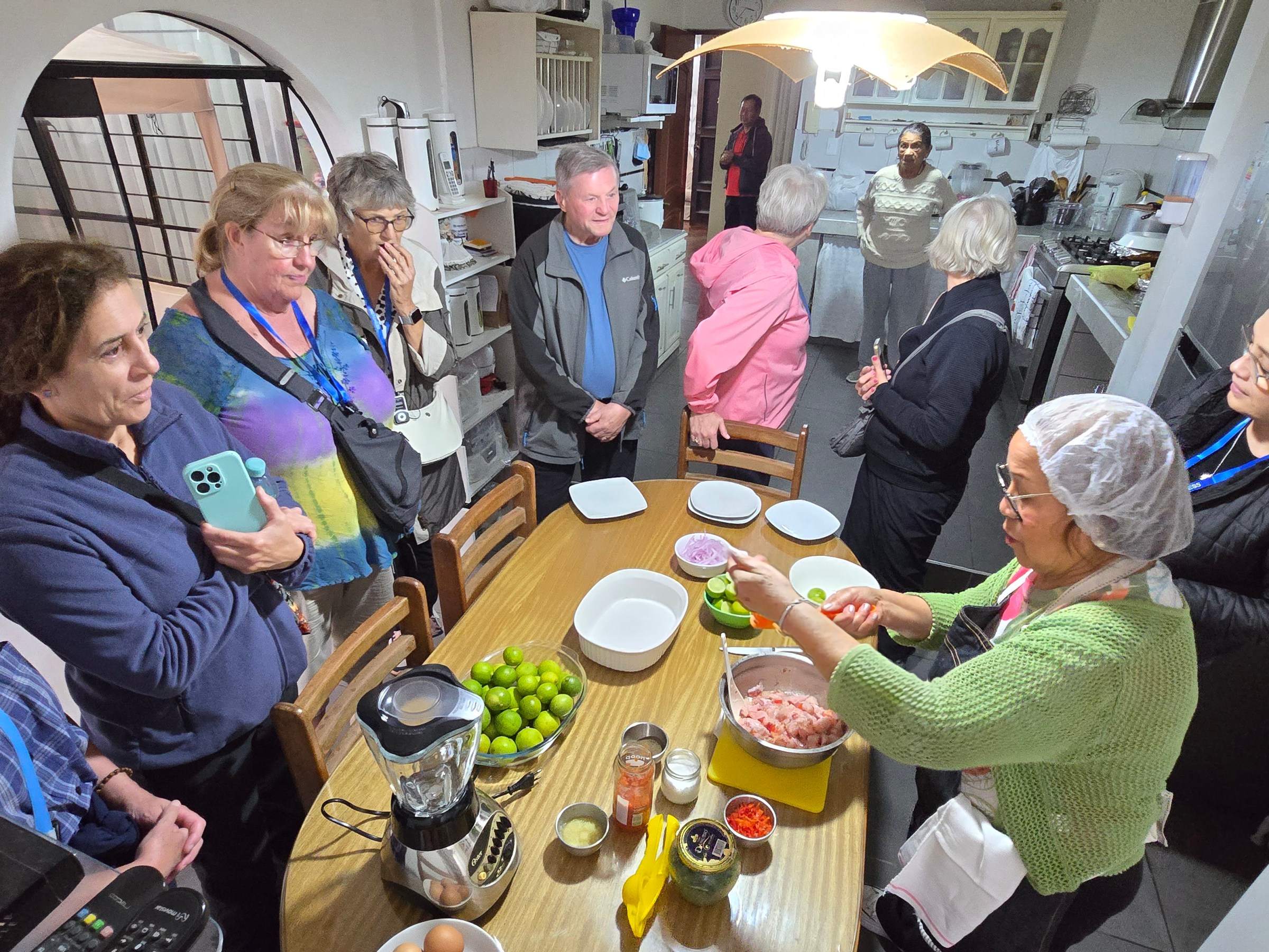 People gather around a kitchen table as a cook demonstrates a recipe with various ingredients.