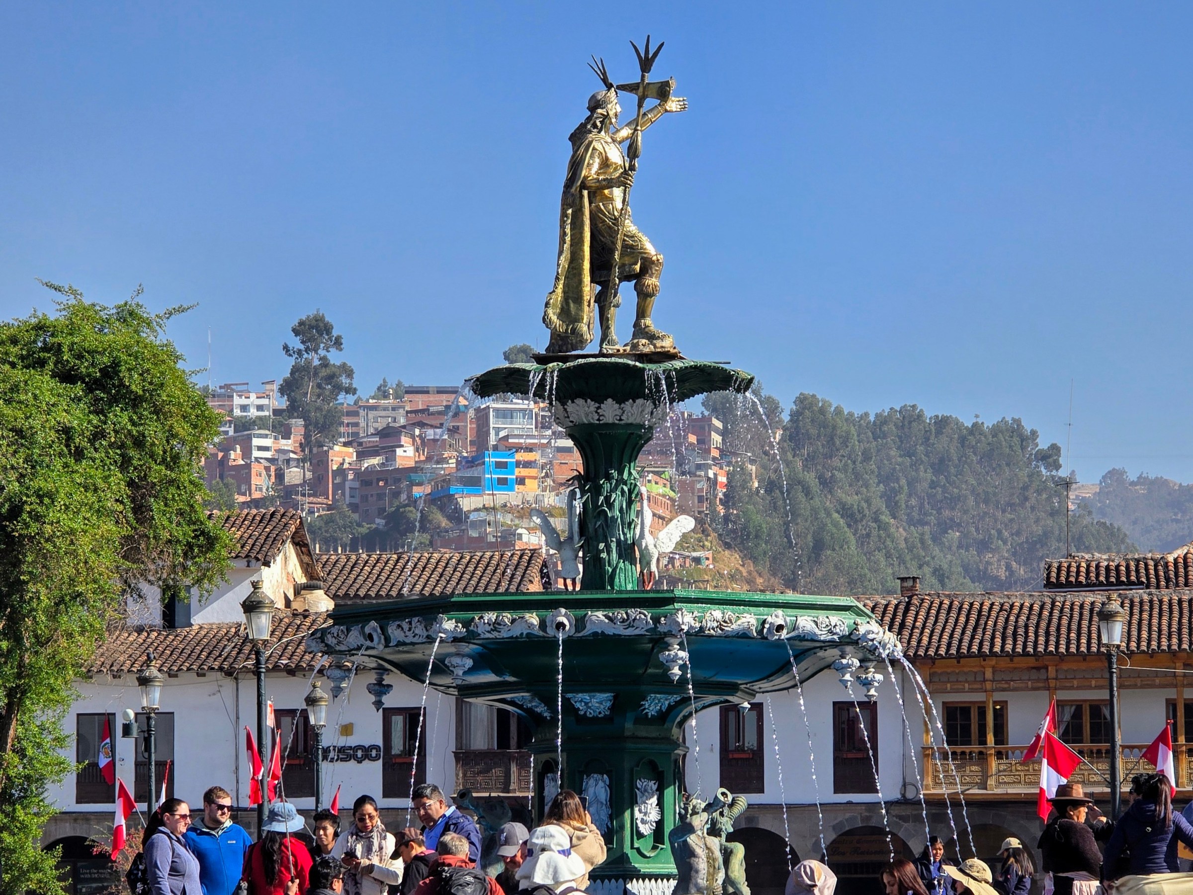 Statue atop a fountain with people around, buildings and hills in background.