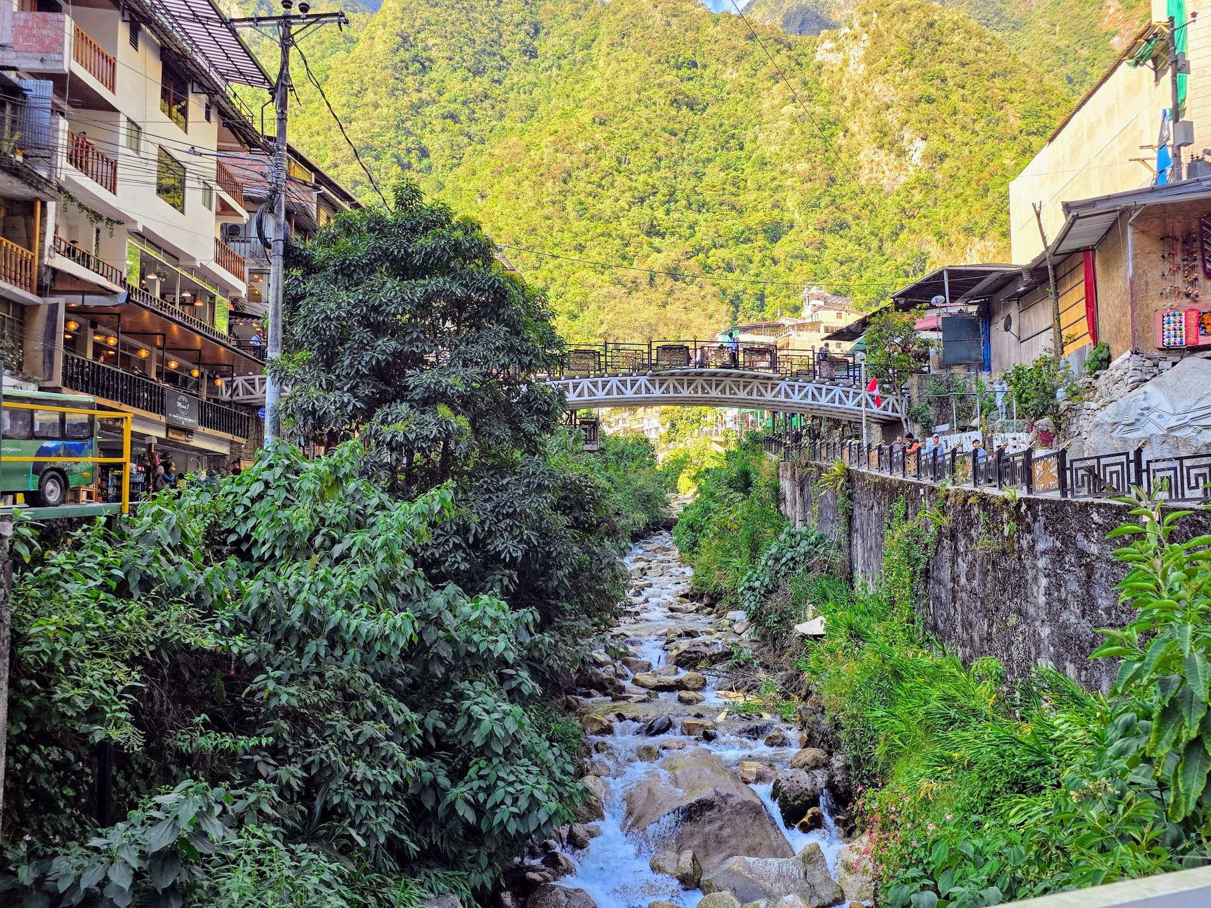 Town buildings beside a flowing river, with a bridge and lush green hillside in the background.